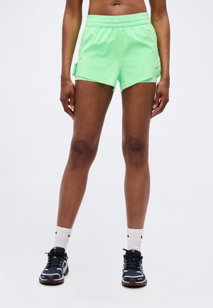 Personne portant un short de sport vert fluo, des chaussettes blanches Nike et des chaussures de course noires, debout devant un fond uni.
