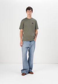 Tricou verde măsliniu cu guler rotund și mâneci scurte, asortat cu blugi largi de culoare albastru deschis. Pantofii maronii completează ținuta.