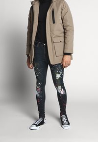 Chaqueta acolchada marrón con un bolsillo con cremallera, suéter negro de cuello alto, jeans de mezclilla desgastados con patrones de salpicaduras y zapatillas altas negras.