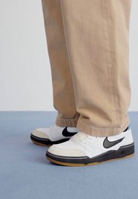 Zapatillas Nike blancas con acentos negros, suela de goma y costuras distintivas. Combinadas con pantalones beiges, mostrando un ajuste relajado.