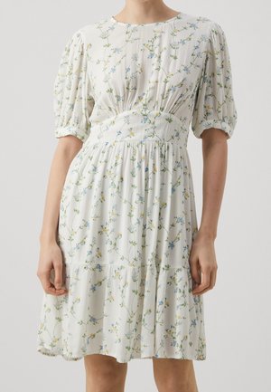Femme portant une robe blanche à motifs floraux bleus et jaunes, manches courtes bouffantes et taille froncée.