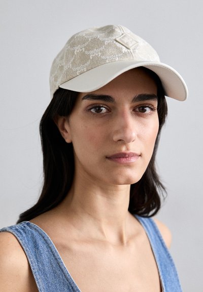 La casquette présente un tissu beige texturé avec une visière blanche, ornée d'une petite étiquette logo à l'avant. Sangle ajustable à l'arrière.