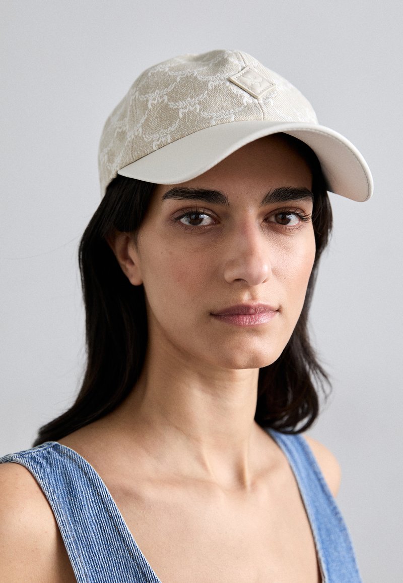 Cap features a textured beige fabric with a white brim, adorned with a small logo patch on the front. Adjustable strap at the back.