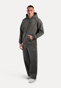 Dark gray zip-up hoodie with a large hood and front pockets, paired with matching sweatpants. Both have a soft, textured finish. White sneakers.