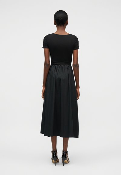 Γυναίκα στέκεται με την πλάτη προς τα εμπρός, φορώντας μαύρο μπλουζάκι με κοντά μανίκια, midi φούστα ψηλόμεση και μαύρα σανδάλια με ψηλό τακούνι σε απλό φόντο.