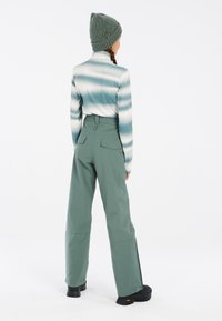 Langsleeve top met blauwe en witte horizontale strepen, gecombineerd met hoge, groene, ruime broek. Model draagt een gestructureerde groene beanie.