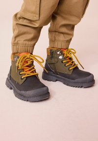 Bottines noires et vertes foncées avec des lacets jaunes et des accents orange. Elles possèdent des semelles texturées et des œillets en métal pour un maintien sécurisé.