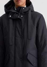 Czarna ocieplana kurtka z kapturem, z napami, bocznymi kieszeniami i detalami z logo na paskach. Gładka tekstura materiału.