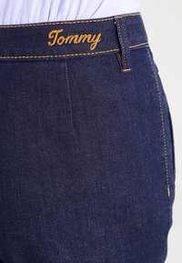 Vaqueros de denim azul oscuro con un logo dorado de "Tommy" en la cinturilla, con costuras de contraste y un corte clásico.