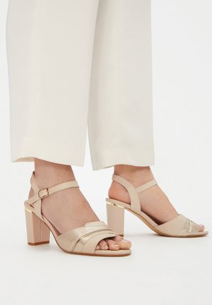 Sandali con tacco beige con dettagli metallici, con una fascia larga sulle dita dei piedi e un cinturino alla caviglia con fibbia. Design con tacco a blocco.