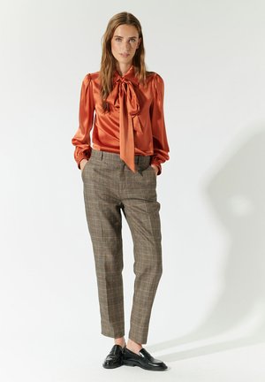 Blusa arancione in satin con fiocco, maniche a sbuffo e pantaloni marroni a quadri. Il modello indossa mocassini neri su uno sfondo neutro.