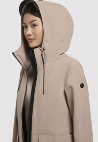 Beige vandtæt jakke med en sort lynlås, justerbar hætte og en glat tekstur. Har et logo på ærmet og forlommer.