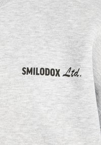 Sweatshirt gris clair avec une texture douce comportant le logo imprimé "SMILODOX Ltd." en noir, positionné sur le côté gauche.