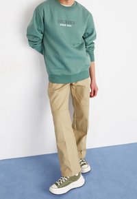 Man i grön Dickies-tröja, beige byxor och gröna låga sneakers, stående på blå matta framför vit vägg.