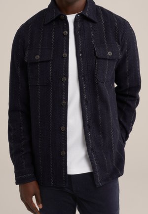 Mann trägt eine marineblaue Jacke mit Knöpfen und vertikalen Streifen über einem weißen T-Shirt, steht mit einer Hand in der Tasche vor einem neutralen Hintergrund.