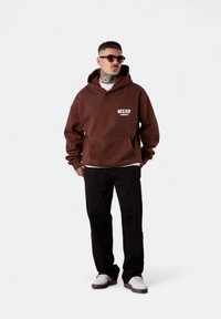 Sweat-shirt à capuche marron avec une poche avant et un texte graphique blanc sur la gauche. Associé à un pantalon noir et des baskets blanches.