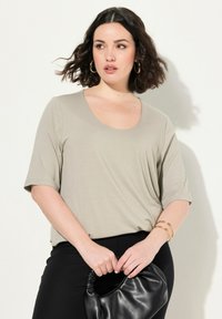 Camiseta de manga corta y escote redondo en un suave tono taupe claro, confeccionada en una tela suave y lisa, combinada con pantalones negros y un bolso negro con fruncidos. Brazaletes dorados en la muñeca.