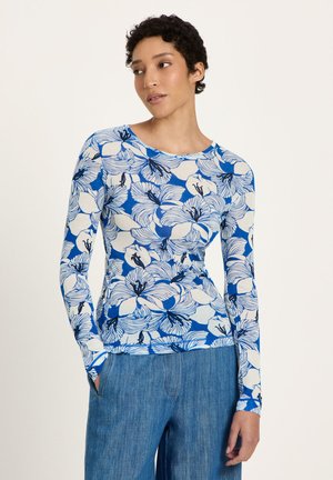 Woman wearing a blue and white floral long-sleeve top and blue jeans, standing with hands in pockets, looking to the side.