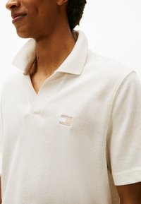 Polo in beige chiaro con una trama testurizzata. Presenta un colletto classico e un piccolo logo ricamato sul lato sinistro del petto. Maniche corte.