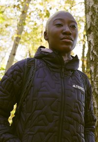 Sort isoleret jakke med et geometrisk quiltet mønster, som har en lynlås og logo. Båret med en rygsæk, sat mod en sløret naturlig baggrund.