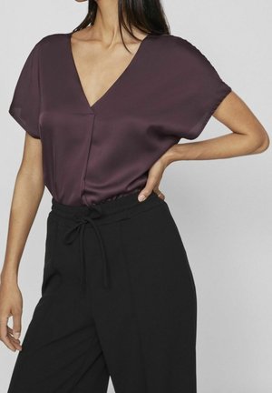 Kobieta w krótkim rękawie fioletowej bluzce z dekoltem w serek, włożonej do wysokich czarnych spodni z suwakiem, pozująca z jedną ręką na biodrze.