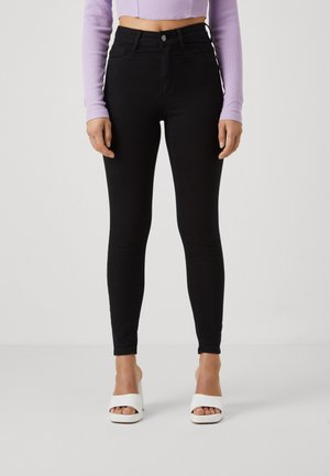 Vrouw draagt zwarte high-waist skinny jeans, een cropped lavendelkleurige top met lange mouwen en witte open-teen sandalen met hak, staand op een effen achtergrond.