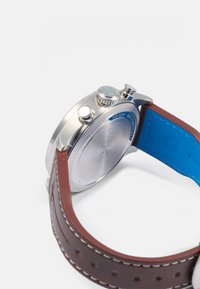 Relógio em aço inoxidável com bracelete de pele marrom e forro azul. Apresenta uma superfície texturizada e dois coroas para ajustes.