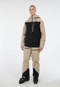 Man die staat in een beige en zwarte skikleding met een capuchonjack, sneeuwbroek en zwarte skischoenen op een witte achtergrond.