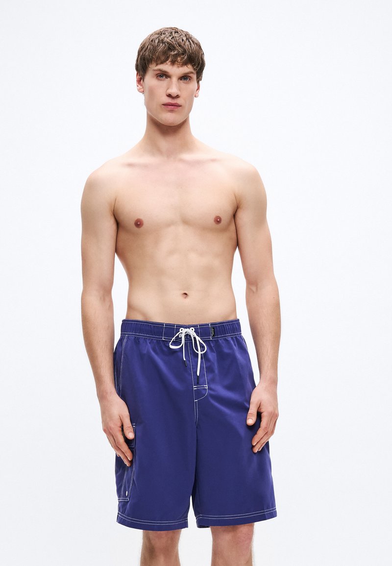 Μωβ μαγιό σορτς με αντίθετη λευκή ραφή, ελαστική μέση και κορδόνι. Διαθέτει πλαϊνή τσέπη και σχεδιασμό με άνετη εφαρμογή.