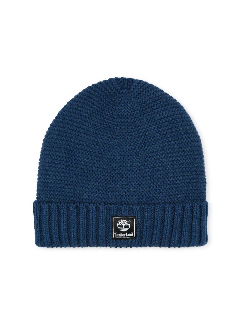 Beanie in maglione di colore blu scuro con risvolto a coste. Presenta una toppa quadrata nera con un logo bianco. Texture morbida e forma arrotondata.