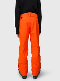 Person iført klare orange vandtætte bukser og sort overdel, stående med ryggen til på en ensfarvet lys baggrund.