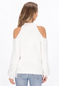 Witte gebreide trui met een cold shoulder ontwerp, hoge hals en franjeranden op de mouwen. Zachte textuur; gedragen met lichtblauwe jeans.