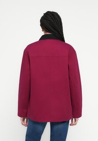 Kurtka w kolorze bordo z czarnym kołnierzem, o luźnym kroju, z długimi rękawami i widocznymi przeszyciami. Noszona na niebieskich dżinsach.