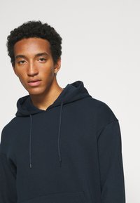 Marineblaue Kapuzenpullover mit Fronttasche, Kordelzügen und entspannter Passform, aus einem weichen Baumwoll-Mischgewebe gefertigt.