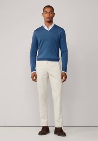 Blå V-ringad tröja, vit skjorta med krage, beige byxor, bruna skor, jämn textur, figursydd design, stående pose mot en neutral bakgrund.