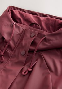 Bordový nepremokavý golier bundy so sťahovacími šnúrkami a hnedými patentkami. Hladký, lesklý povrch s zaobleným tvarom a vnútornou podšívkou.