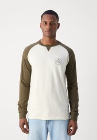 Pika pikkade varrukatega T-särk heledast kreemivärvi kehast ja tumerohelistest raglanvarrukatest. Rinnas logo ja pehme puuvillane materjal.