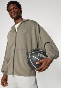 Grå zip-up hættejakke med "Los Angeles Lakers" logo, løs pasform, kengurulommer og ribbede manchetter. Model holder en sort basketball.
