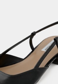 Fekete bőr pántos cipő hegyes orral, vékony pántokkal és alacsony blokk sarokkal. Sima textúrájú, minimalista dizájnelemekkel.