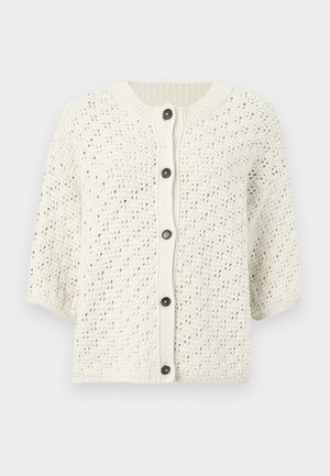 Knit cardigan in cream with open weave and black buttons, paired with light grey wide-leg denim pants. Model wearing cream shoes.