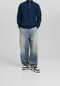 Sudadera de cuello con cremallera en navy, corte relajado. Jeans sueltos en azul claro con textura desgastada. Calzado con acentos en blanco y negro.
