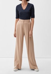 Nainen, joka on pukeutunut laivansinisessä V-pääntiellä varustettuun yläosaan, jossa on kyynärpäihin ulottuvat hihat, beigeihin korkeavyötäröisiin leveälahkeisiin housuihin ja beigeihin lenkkareihin, seisoo valkoisella lattialla.