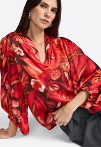 Kvinde med langt m�ørkt hår iført sølvøreringe og en rød bluse med blomsterprint og voluminøse ærmer, siddende på en hvid overflade.
