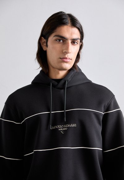 Jovem com cabelo escuro até aos ombros, vestindo um hoodie preto da Emporio Armani com detalhes brancos e logótipo, sobre um fundo claro e liso.