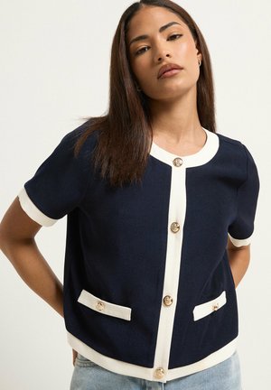 Kobieta w granatowym krótkim kardiganie z białym wykończeniem i złotymi guzikami, w połączeniu z jasnoniebieskimi jeansami, na jednolitym tle.