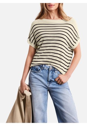 Kobieta ubrana w beżowy i czarny pasiasty sweter z krótkim rękawem oraz jasnoniebieskie dżinsy, trzymająca beżową kurtkę w jednej ręce.