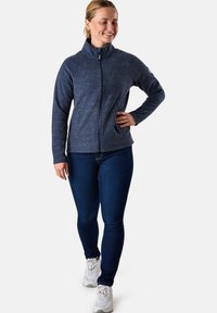 Giacca in pile con zip di colore blu navy, con colletto alto, design aderente e tessuto a trama; abbinata a jeans blu scuro e sneakers bianche.