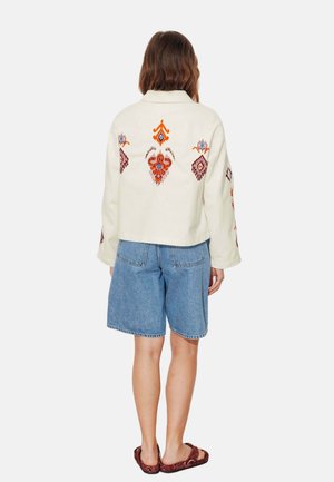 Femme portant une veste crème avec une broderie tribale colorée, un short en jean bleu et des sandales rouges à motifs, debout de dos.