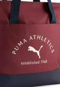 Punainen ja laivastonsininen olkusäkkijuttu, jossa on teksturoidusta kankaasta valmistettu pinta, valkoinen "Puma Athletics" -logo ja perustamisvuosi, vahvistetut olkahihnat.