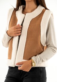 Gilet in suede finto beige con bordi in pelliccia finta bianca, dotato di scollatura rotonda e senza chiusure, indossato sopra una maglietta grigia a maniche lunghe.